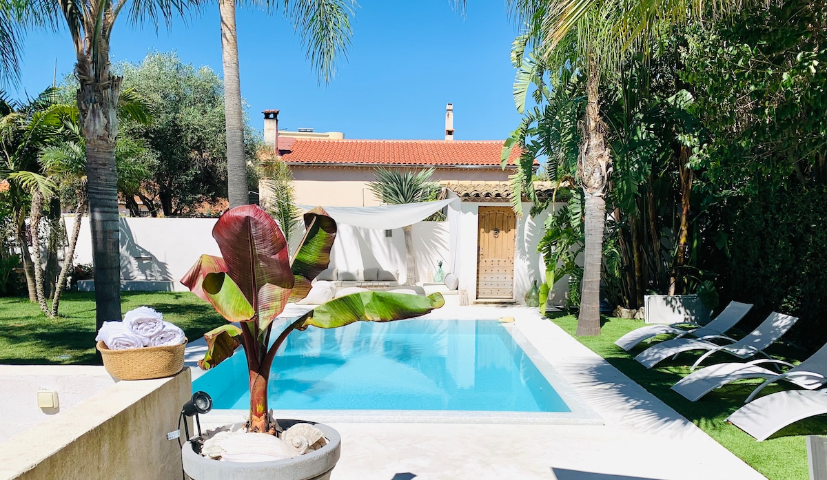 A serene garden area showcases a clear swimming pool surrounded by lounge chairs. Tall palm trees and lush greenery frame the space, creating a tranquil environment. Soft towels are neatly arranged in a nearby bowl, enhancing the relaxing atmosphere of the outdoor area.