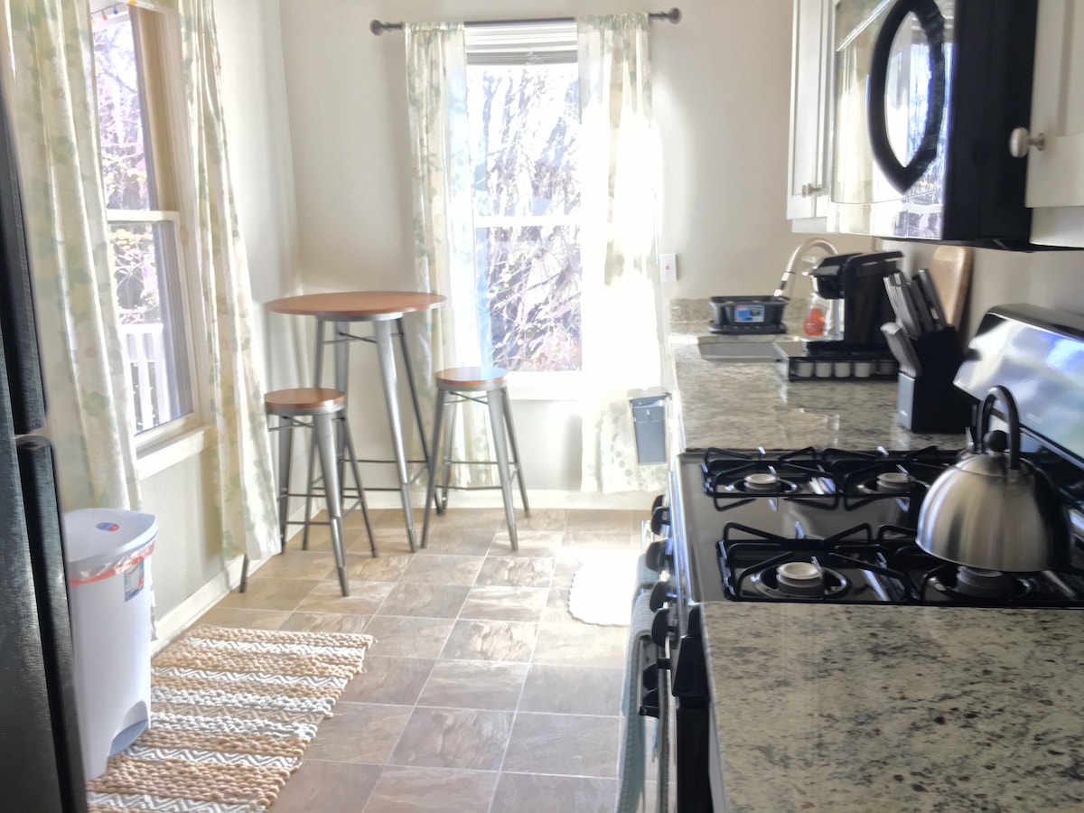 A kitchen is illuminated by natural light through two windows, with bar stools positioned around a high table. The countertop features a stove, kettle, and various kitchen appliances, complemented by a woven rug on the floor. Neutral colors enhance the overall space.