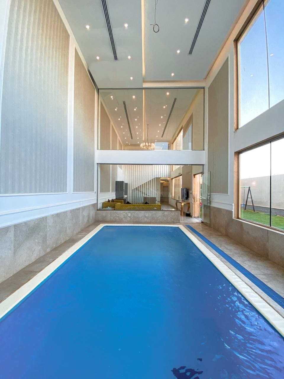 An indoor swimming pool is depicted, featuring clear blue water and a smooth tile surface. Tall windows allow natural light to illuminate the space, while elegant lighting fixtures enhance the atmosphere. A view of the adjacent lounge area and garden can be seen in the background.