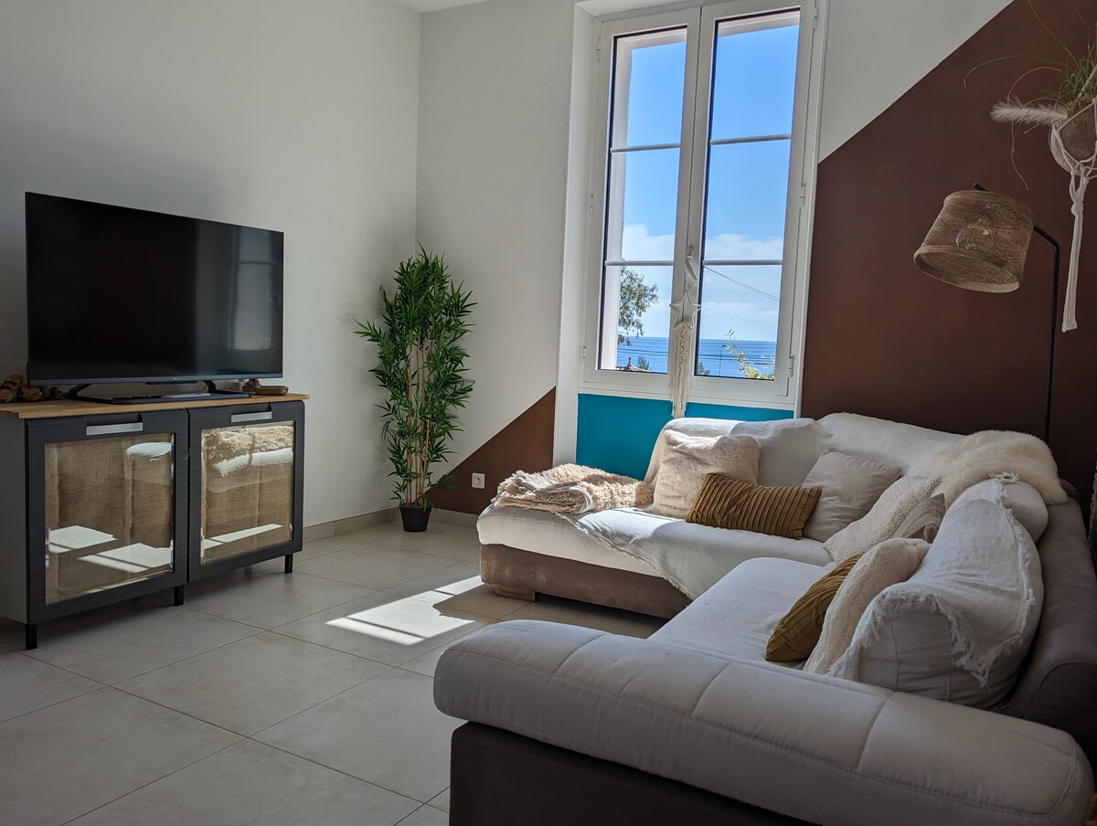 A spacious living area is presented, featuring a corner sectional sofa and a flat-screen television. Natural light enters through large windows that offer a view of the sea. A potted plant adds a touch of greenery to the modern design.