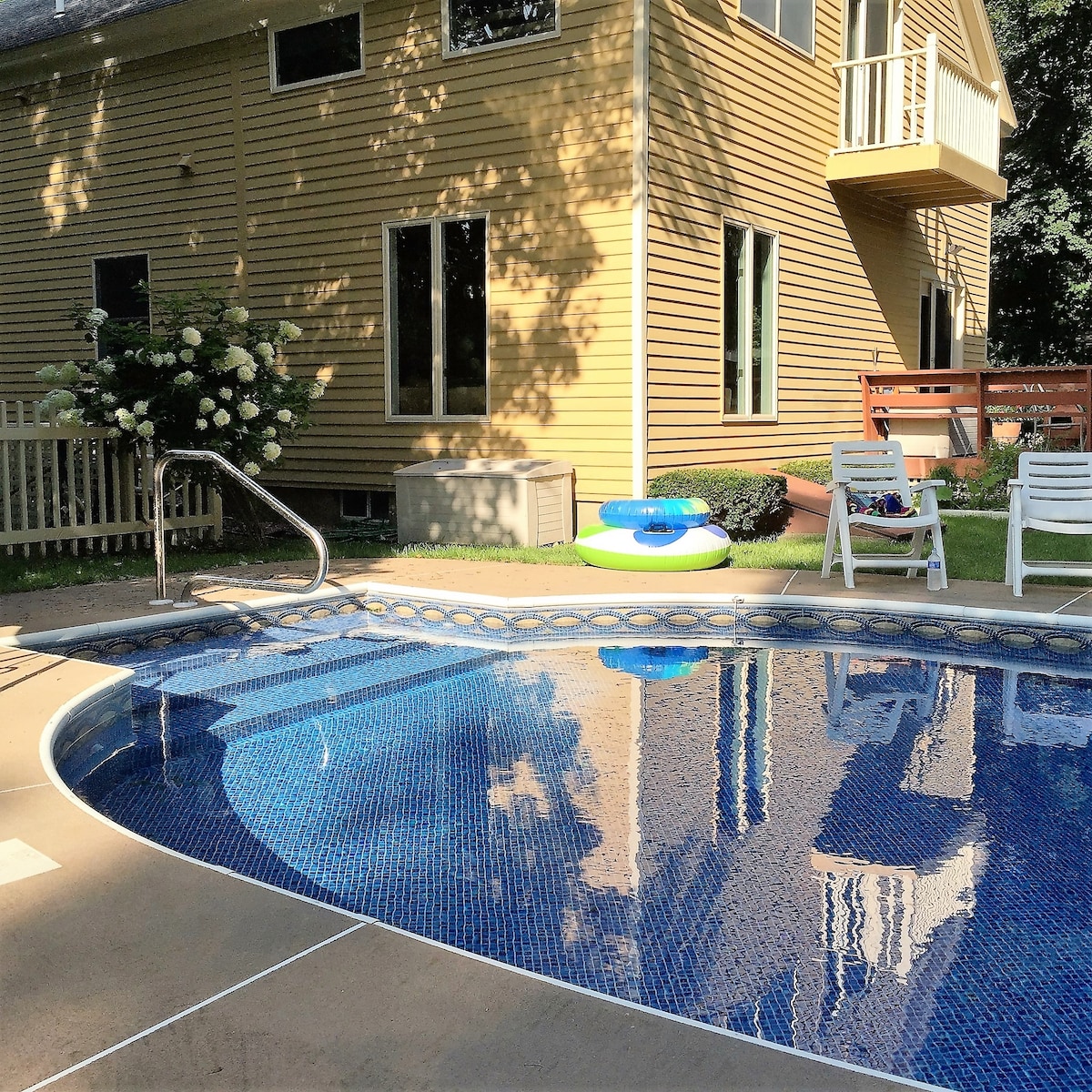 An inground pool is surrounded by a sunlit patio featuring a colorful inflatable float and white folding chairs. A yellow house with large windows, framed by green trees, is visible in the background, showcasing a blend of indoor and outdoor spaces.