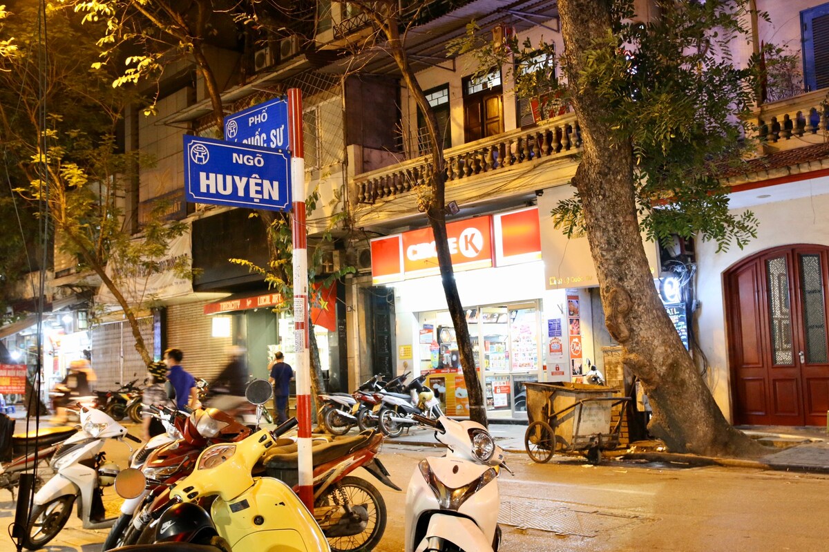 The street view captures a lively urban scene, with the corner street sign indicating 'Huyen.' A convenience store and parked motorcycles are visible, framed by trees that provide a natural element to the busy environment. Soft evening lighting enhances the neighborhood's welcoming atmosphere.