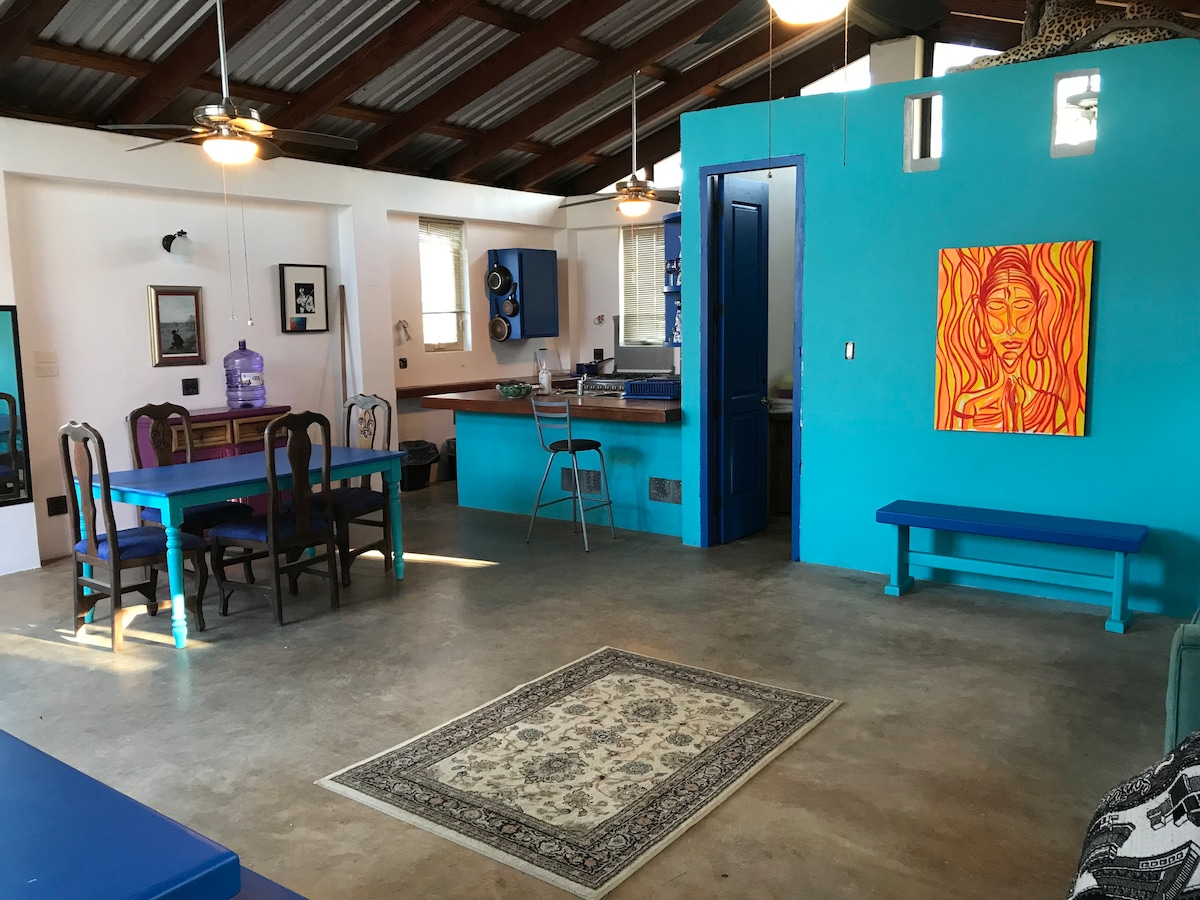 A spacious living area features vibrant blue walls and a high ceiling with wood beams. A dining table with colorful chairs is positioned near a fully equipped kitchen. Illuminated by natural light, artwork decorates the walls, and a decorative rug lies on the concrete floor.