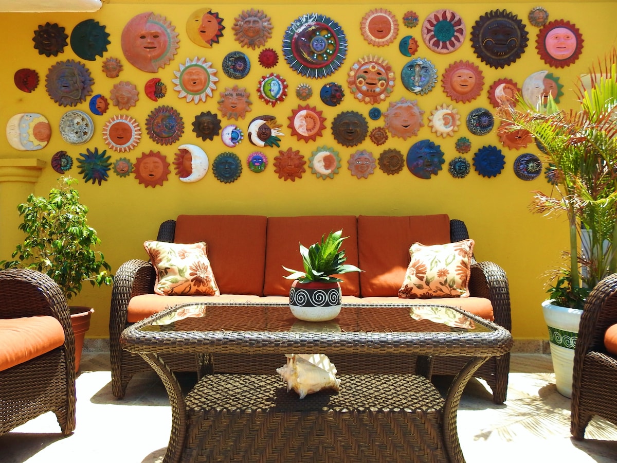 A vibrant seating area features a brown wicker sofa adorned with orange cushions, positioned in front of a wall decorated with an array of colorful sun-themed wall art. A glass coffee table sits in the center, topped with a decorative shell and a potted plant.