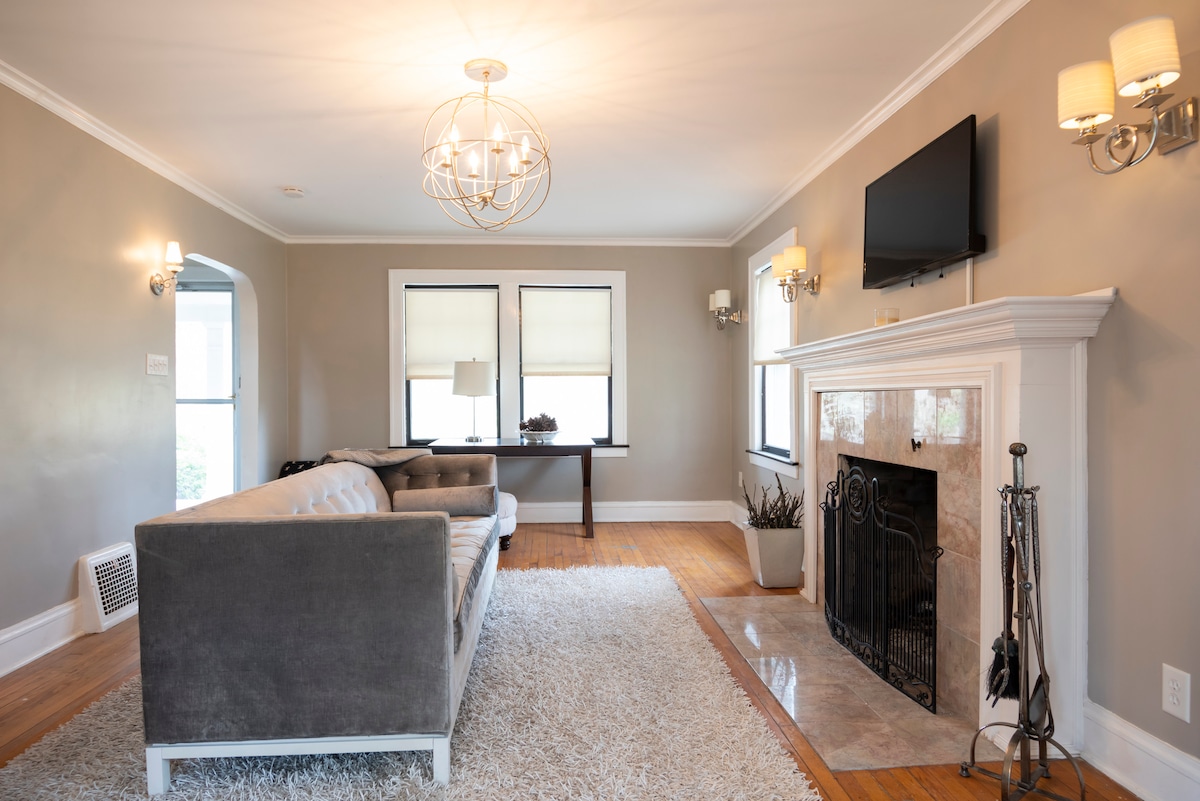 A spacious living room exudes comfort with a large gray sectional sofa and a soft area rug. Natural light fills the space through two windows. A modern chandelier and wall sconces provide ample lighting, while a decorative fireplace anchors the room beneath a mounted television.