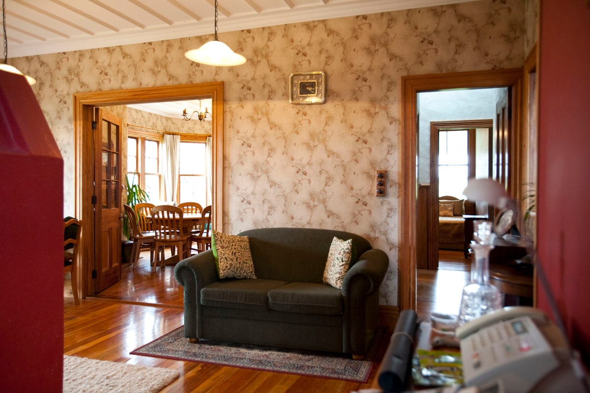 A cozy living area features a green sofa with decorative cushions, positioned on a hardwood floor. Light fixtures hang from the ceiling, illuminating floral-patterned wallpaper. Doorways lead to additional rooms, including a dining area with wooden chairs and a nearby cozy sitting room.