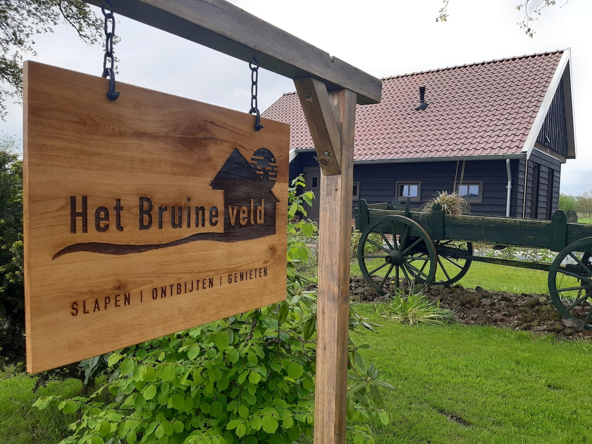 A wooden sign featuring the text 'Het Bruine veld' hangs prominently, surrounded by green foliage. In the background, a dark wood building with a red roof is visible, along with an antique wooden cart resting on the grass.