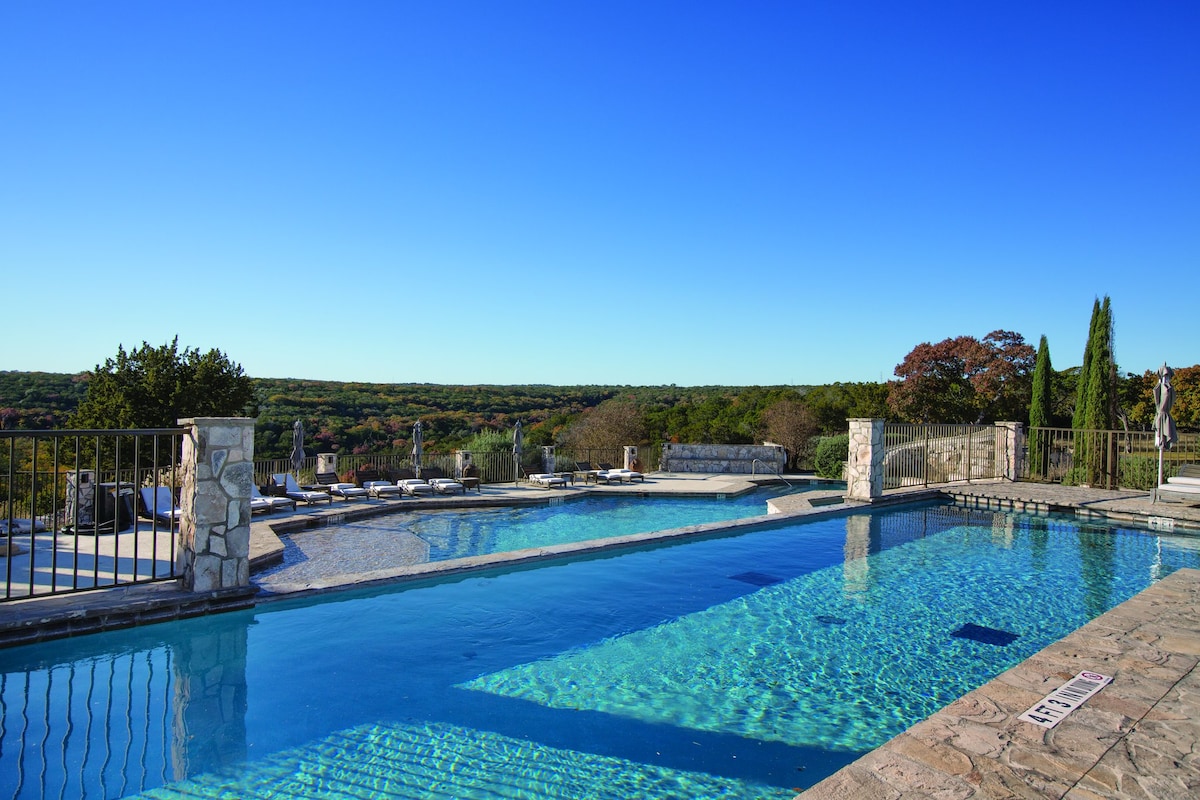 Two outdoor pools are visible, featuring clear blue water and surrounded by natural stone. A railing encloses the pools, offering scenic views of the green hills in the background under a clear blue sky.