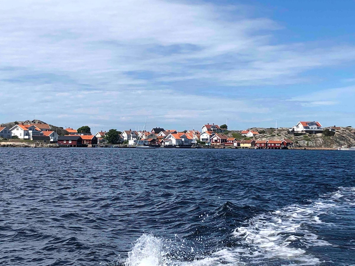 A scenic view of a coastal village is captured from the water, showcasing traditional houses with orange roofs lining the shoreline. The calm blue water reflects patches of sky, while rocky cliffs are visible in the background, contributing to the natural beauty of the area.