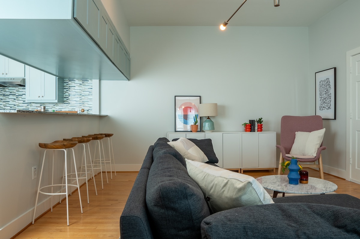 A modern living space features a comfortable dark sofa adorned with several cushions. A round coffee table is positioned in front, while a light-colored chair adds contrast. The kitchen area is visible in the background, complemented by sleek bar stools. Large windows allow natural light to fill the room.
