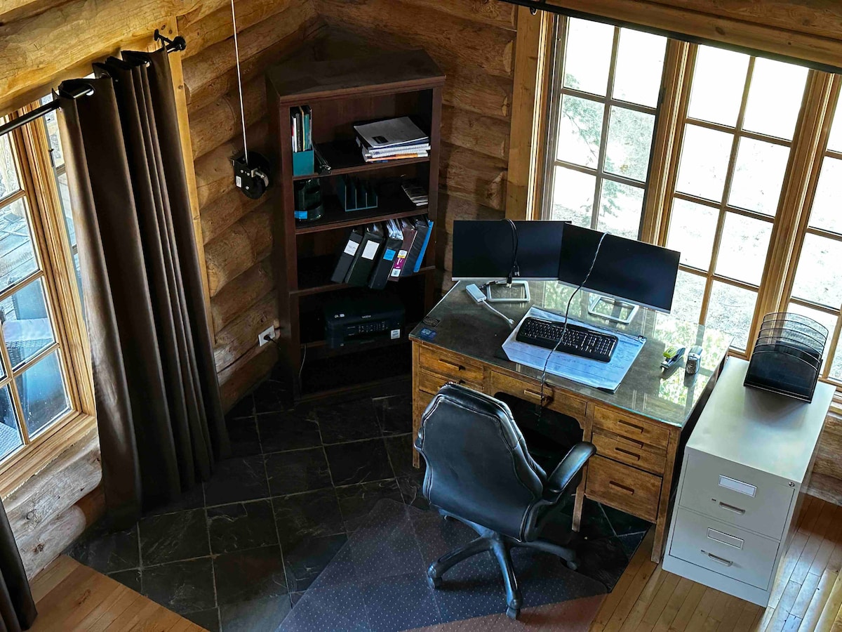 An organized workspace is highlighted within a wooden cabin. A sleek desk hosts dual monitors and various office supplies. Natural light floods the area through large windows, while dark curtains add a touch of privacy. The flooring features a mix of dark tiles and wooden elements.