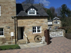 The exterior of Keepers Cottage is highlighted, featuring natural stone walls and a pitched roof. A decorative wooden wheel is positioned next to the entrance, and a small white chair is visible. The cobblestone driveway leads to a fenced area, providing access to the property.