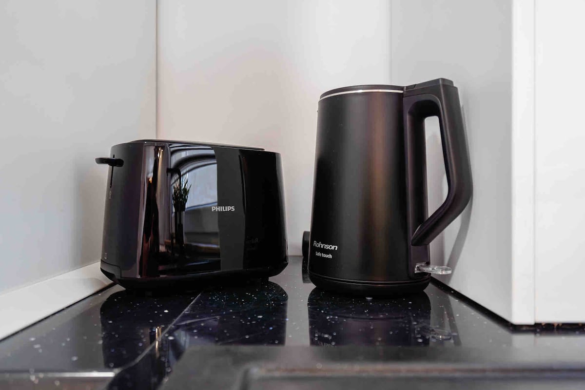 A sleek black toaster and a black electric kettle are positioned on a dark countertop. The simple design of the appliances complements the modern kitchen space, with a hint of natural light visible reflecting off the countertop.