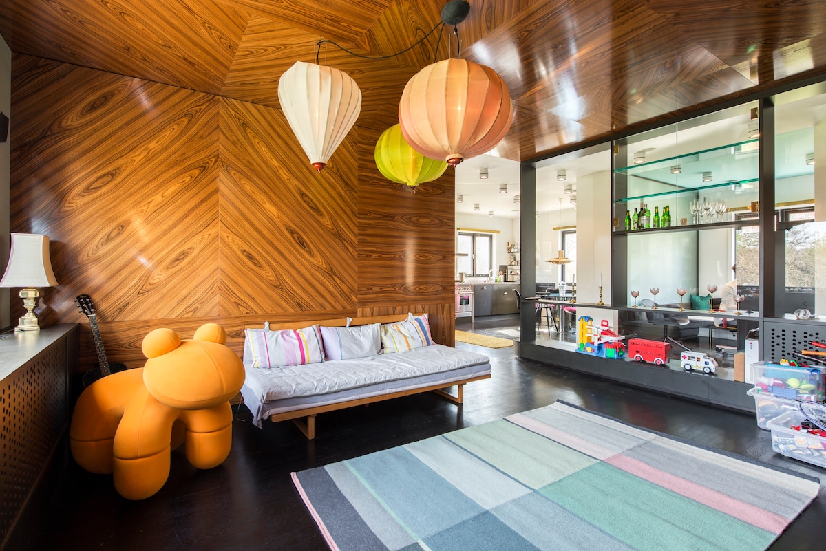 A warm-toned wooden wall features a geometric design, complemented by colorful hanging lanterns. A low seating area is positioned against the wall, adorned with multi-colored cushions. A soft, striped rug lies beneath, while a play area with toys is visible in the corner.