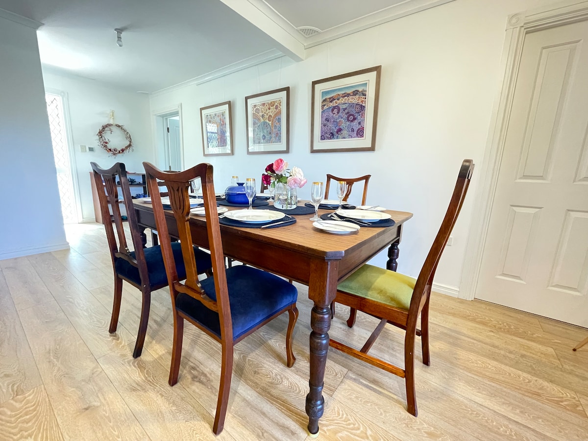 A dining area is shown with a polished wooden table set for a meal, featuring an arrangement of fresh flowers. Surrounding chairs in various colors provide seating, while light from a nearby window brightens the space. Artwork adorns the walls, enhancing the ambiance.