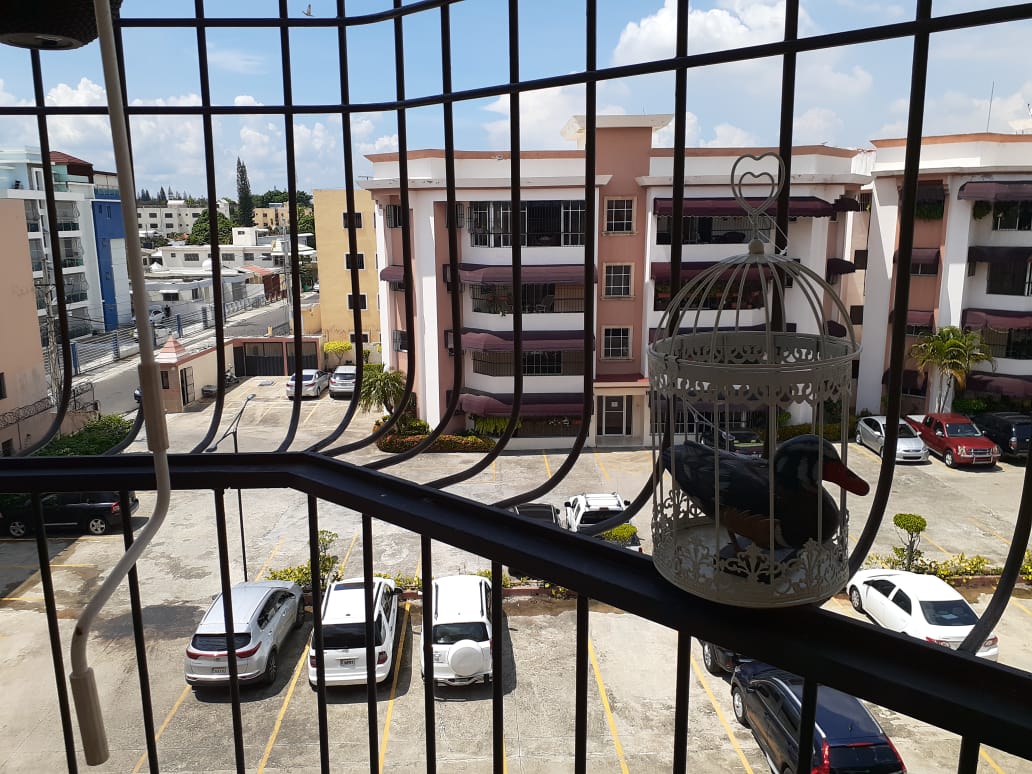 A view from the balcony shows a parking area with several cars and a well-maintained landscape. Decorative metalwork of a birdcage is visible in the foreground, contrasting with the residential buildings and blue sky in the background.