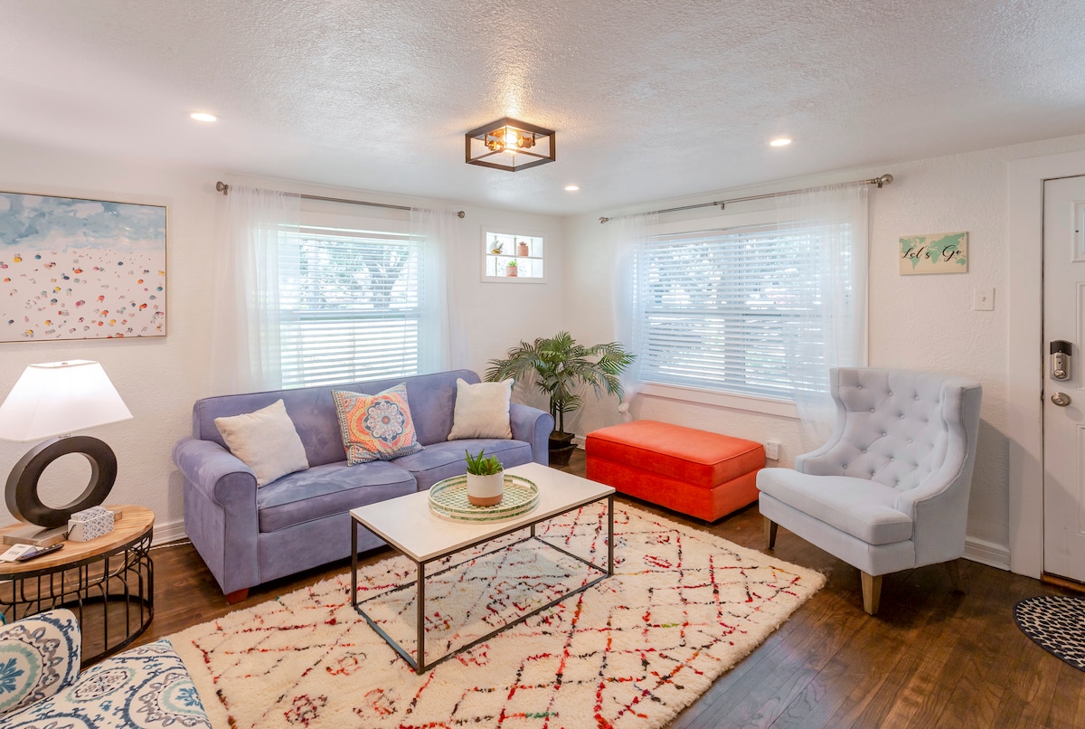 A bright living area features a lavender sofa and a coral ottoman, complemented by a light blue armchair. Natural light fills the space through multiple windows, with a small indoor plant accentuating the coffee table. A patterned area rug adds texture to the hardwood floor.