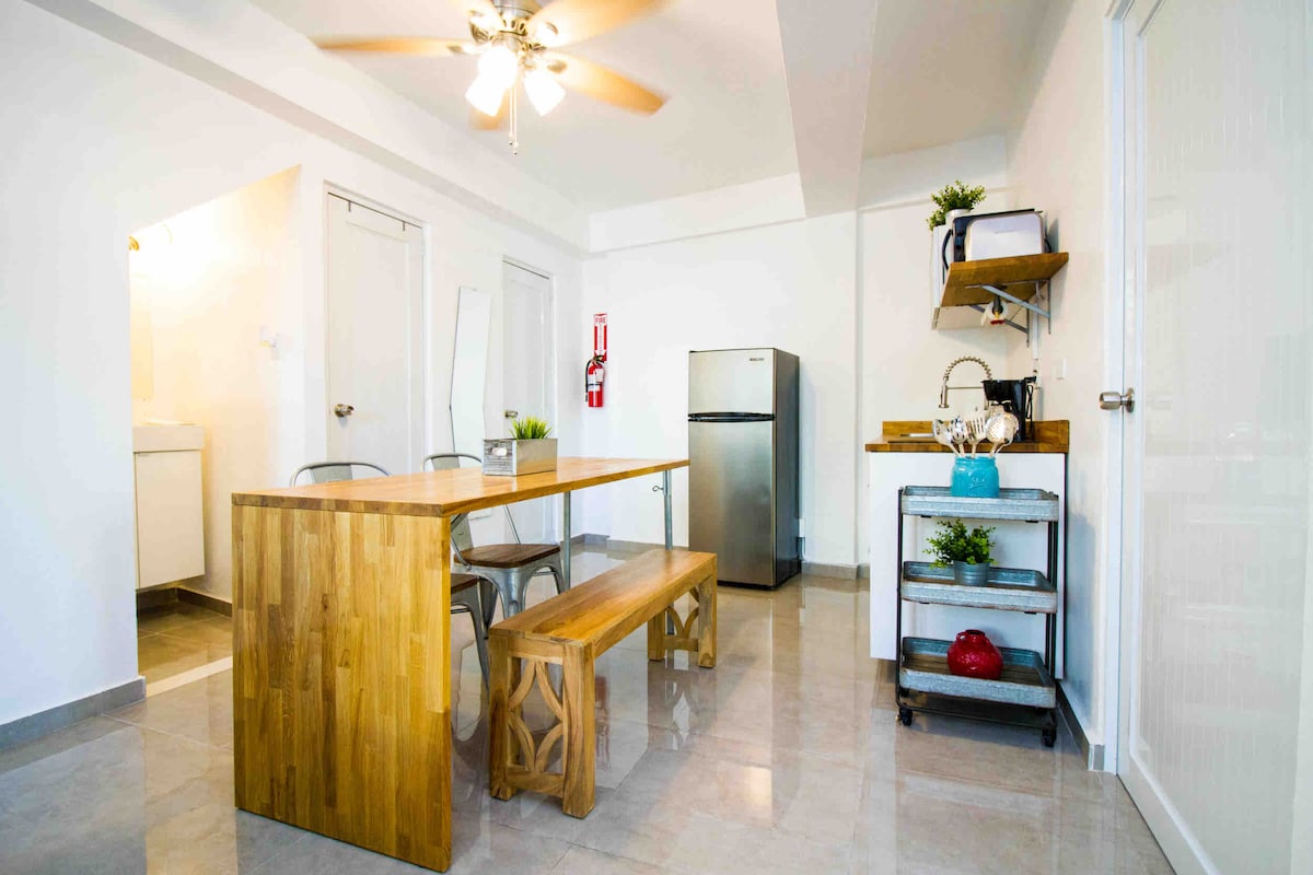 A bright dining area features a wooden table paired with a matching bench and metal chairs. A small cart hosts kitchen supplies, while a refrigerator is positioned nearby. A ceiling fan hangs above, offering ventilation in the open space. Natural light supplements the modern aesthetic.