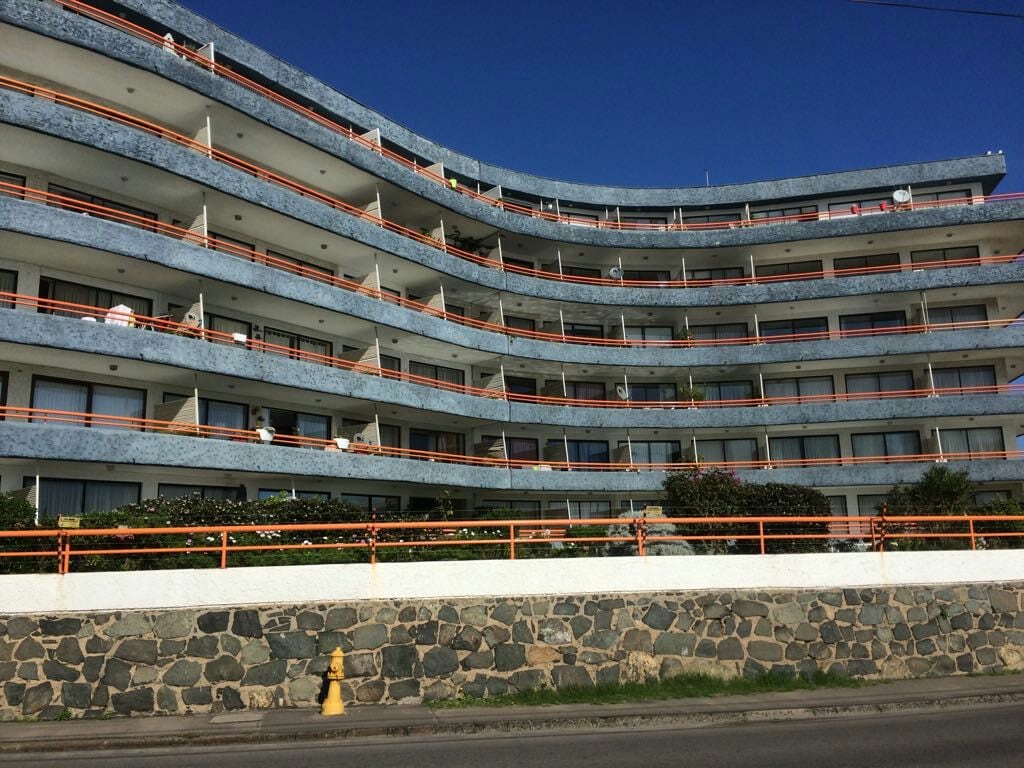 The exterior of a multi-story building is displayed, featuring a modern design with multiple balconies lined with orange railings. Lush greenery is visible at the base, and blue skies create a bright backdrop. The structure provides direct access to the beach.