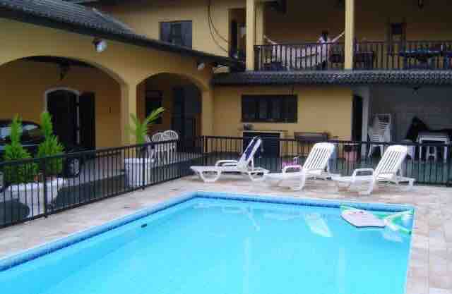 A well-maintained pool area is shown, featuring a clear blue water pool surrounded by several lounge chairs. The patio area includes a shaded balcony and various outdoor furniture, with scattered potted plants adding a touch of greenery.