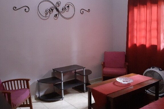A small living area is captured, featuring two pink armchairs and a wooden coffee table. A dark shelving unit stands against the wall, with natural light filtering through a large window draped in red curtains. Decorative wall art enhances the space.