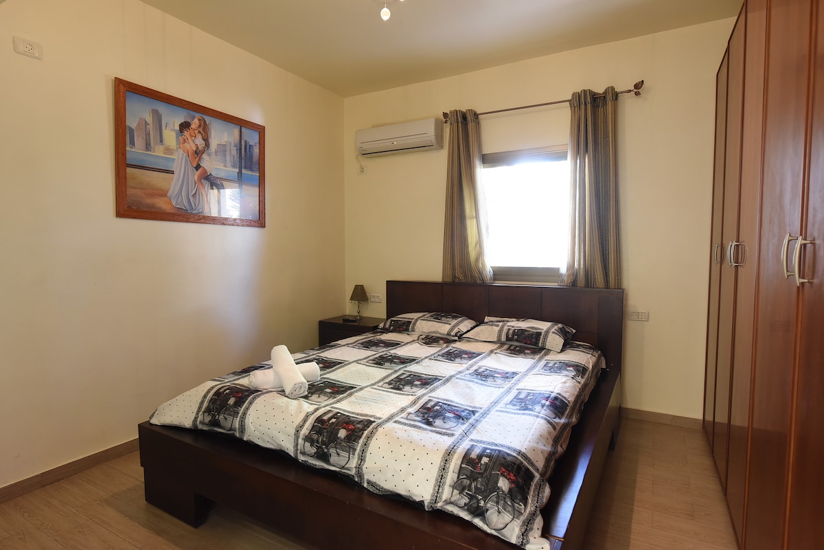A double room features a spacious bed dressed in a patterned blanket. Natural light enters through a window covered by light curtains, and a piece of artwork is displayed on the wall. Built-in wardrobes provide ample storage space.