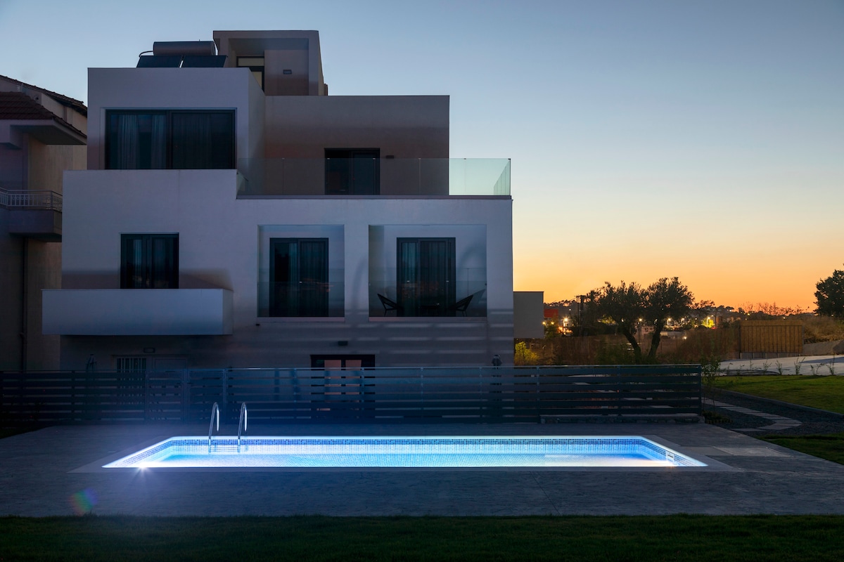 A modern building is showcased against a twilight sky, with a well-lit swimming pool in the foreground. The pool features blue illumination, creating a calming effect. The structure has multiple large glass windows, allowing glimpses of the interiors and potential outdoor seating.