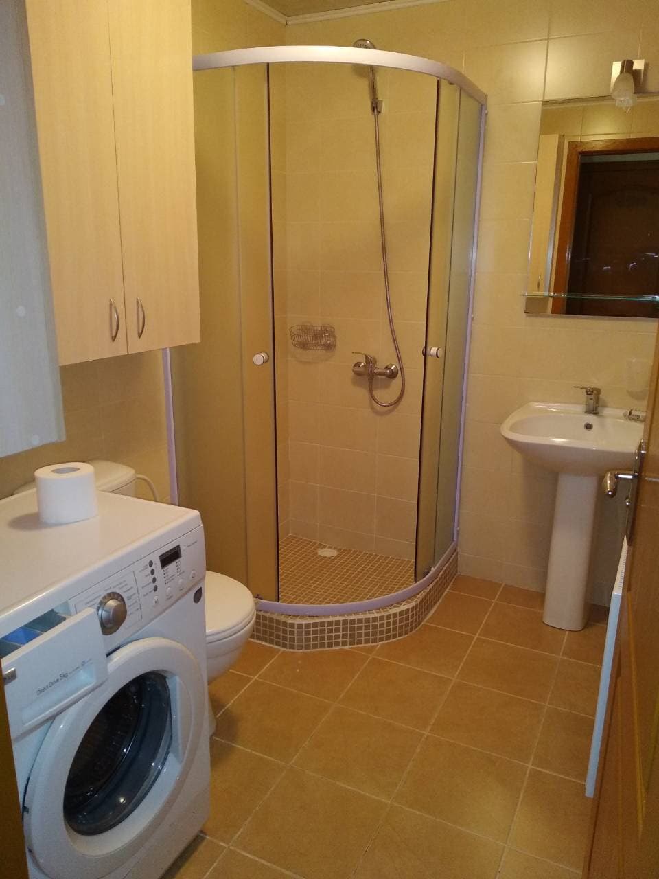 A functional bathroom is equipped with a shower enclosure and a modern sink. A washing machine is situated beside a toilet, and cabinetry provides storage. Neutral tones dominate the space, designed for practicality and comfort.
