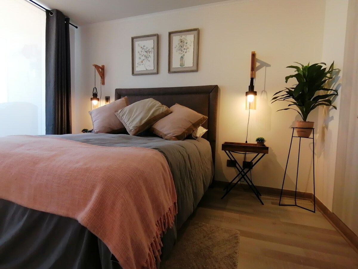 A cozy sleeping area is presented with a spacious bed dressed in neutral linens. Two decorative wall lamps provide soft lighting, with floral art pieces adorning the walls. A potted plant and a small side table complement the room's tranquil aesthetic.
