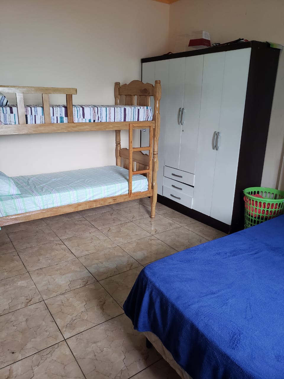 A cozy room features a bunk bed with a light green and white checkered pattern and a spacious bed covered with a blue blanket. A wardrobe in contrasting black and white color stands against the wall, complemented by a green basket nearby.