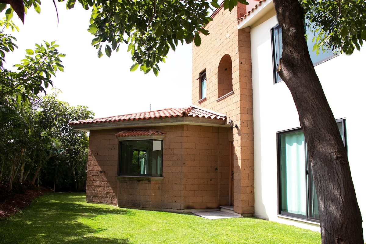 The exterior of a modern two-story house is viewed from the garden. Natural stone and light-colored walls are complemented by a tiled roof. A spacious lawn with greenery surrounds the structure, while a tree offers shade nearby.