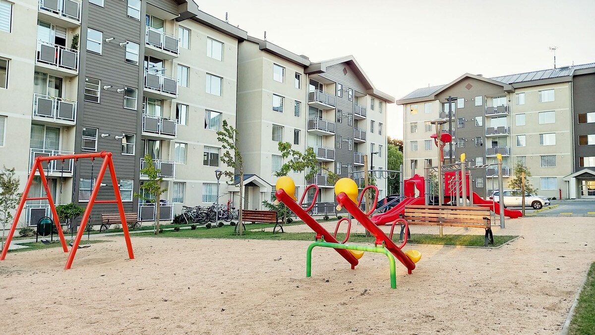 A playground area is located in an open space surrounded by buildings. Colorful swings and a seesaw are present, along with bright play structures. Soft sand covers the ground, offering a safe surface for children. Green trees and benches provide a comforting setting.