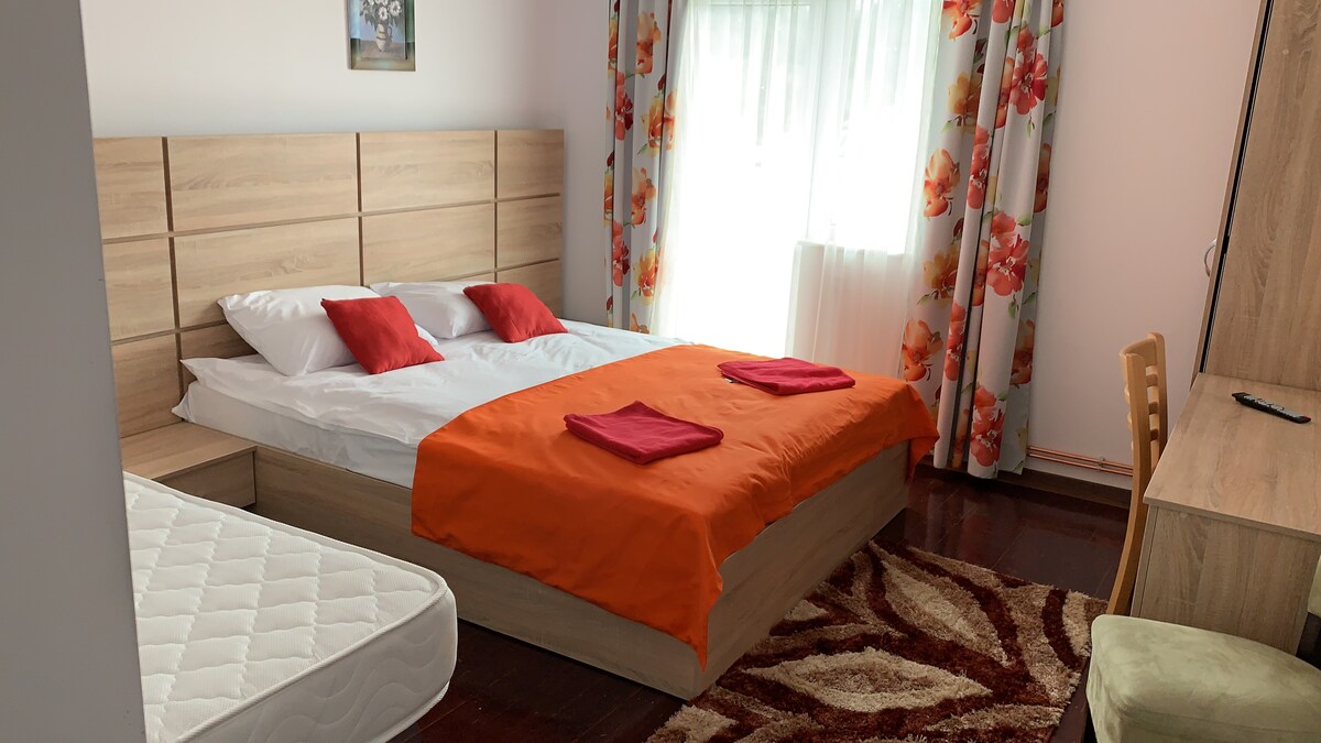 A spacious sleeping area features a large bed with an orange blanket and matching towels. Decorative floral curtains filter natural light, while a desk and a cozy chair are positioned nearby. A partial view of a mattress is seen beside the bed.