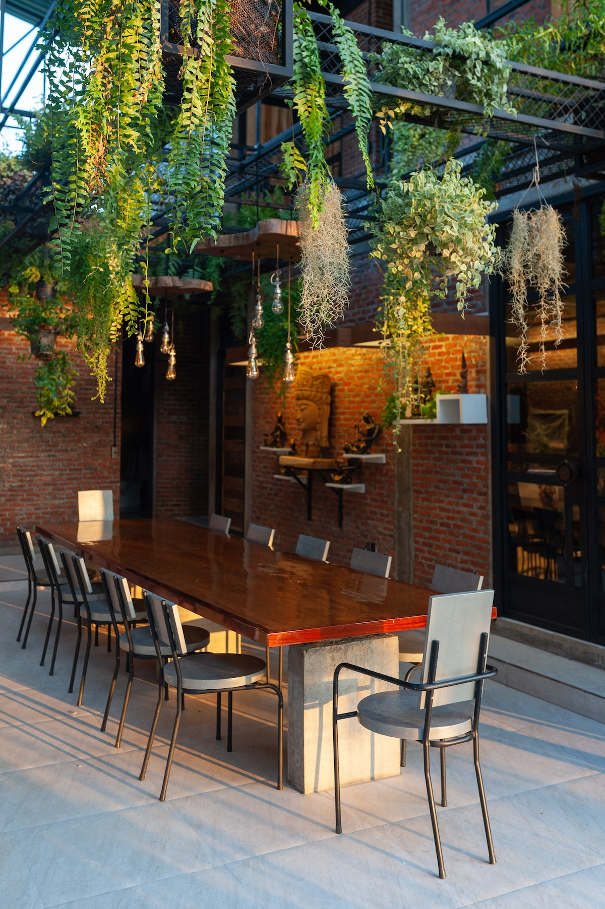 A spacious outdoor dining area is outlined by hanging plants and features a long wooden table surrounded by eight chairs. The brick walls and natural light create a serene setting for communal meals.
