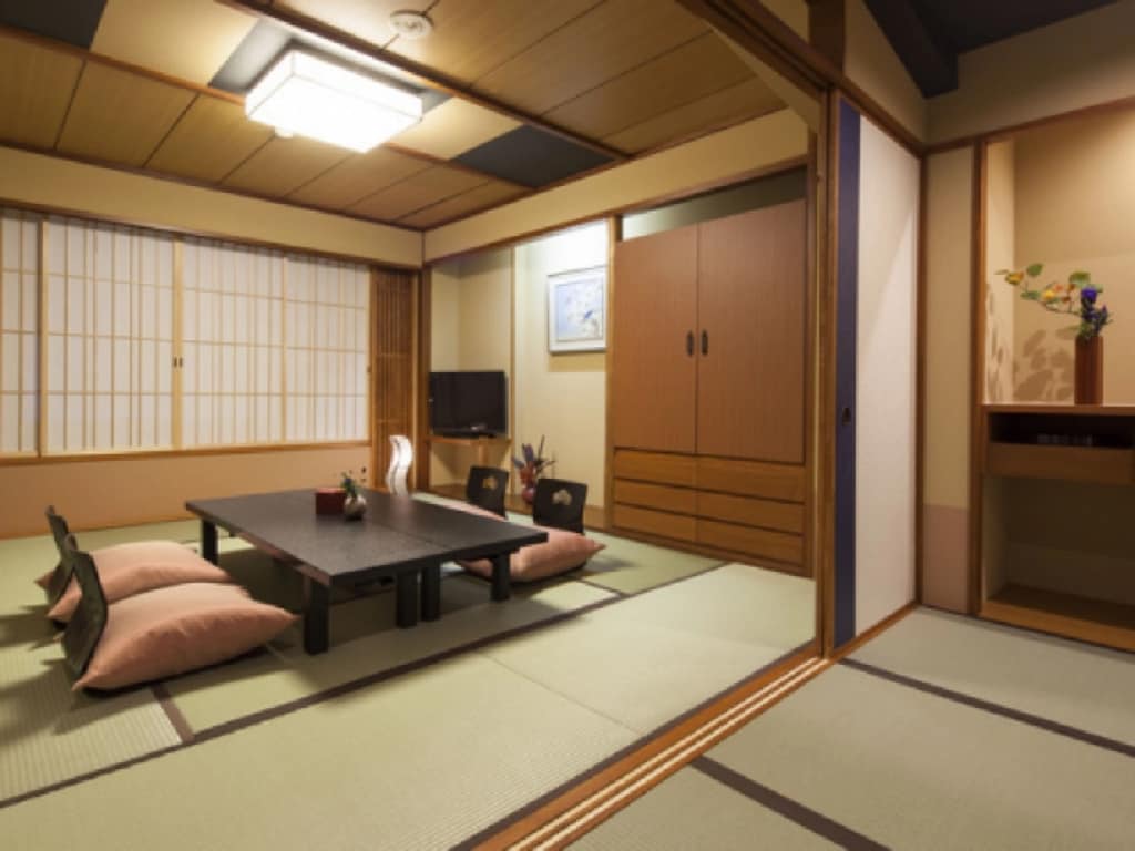 A modern Japanese-style room is presented with tatami mat flooring and sliding shoji doors. A low black table is surrounded by black chairs and cushions. A mounted flat-screen TV is visible along with wooden cabinetry and a small floral arrangement on display.