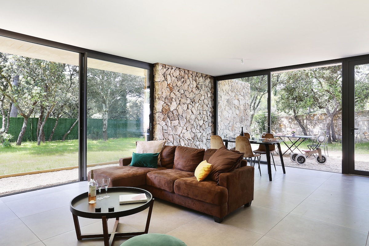 A spacious living area is surrounded by large glass walls, allowing natural light to fill the space. A brown sectional sofa, accented with colorful pillows, is positioned near a low coffee table. A stone feature wall enhances the contemporary design, with garden views visible through the glass.