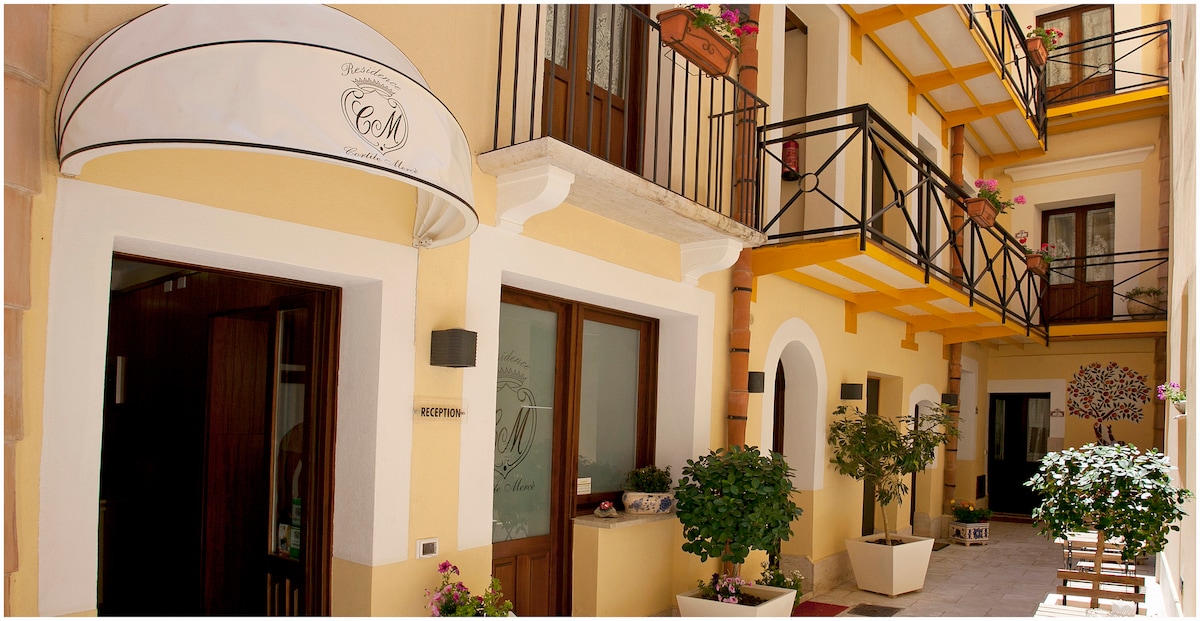 A sunlit courtyard features yellow walls and multiple flower planters. Balconies with black railings overlook the area, which is decorated with greenery. A covered entrance is visible, leading into the building. The space appears inviting and perfect for relaxation.