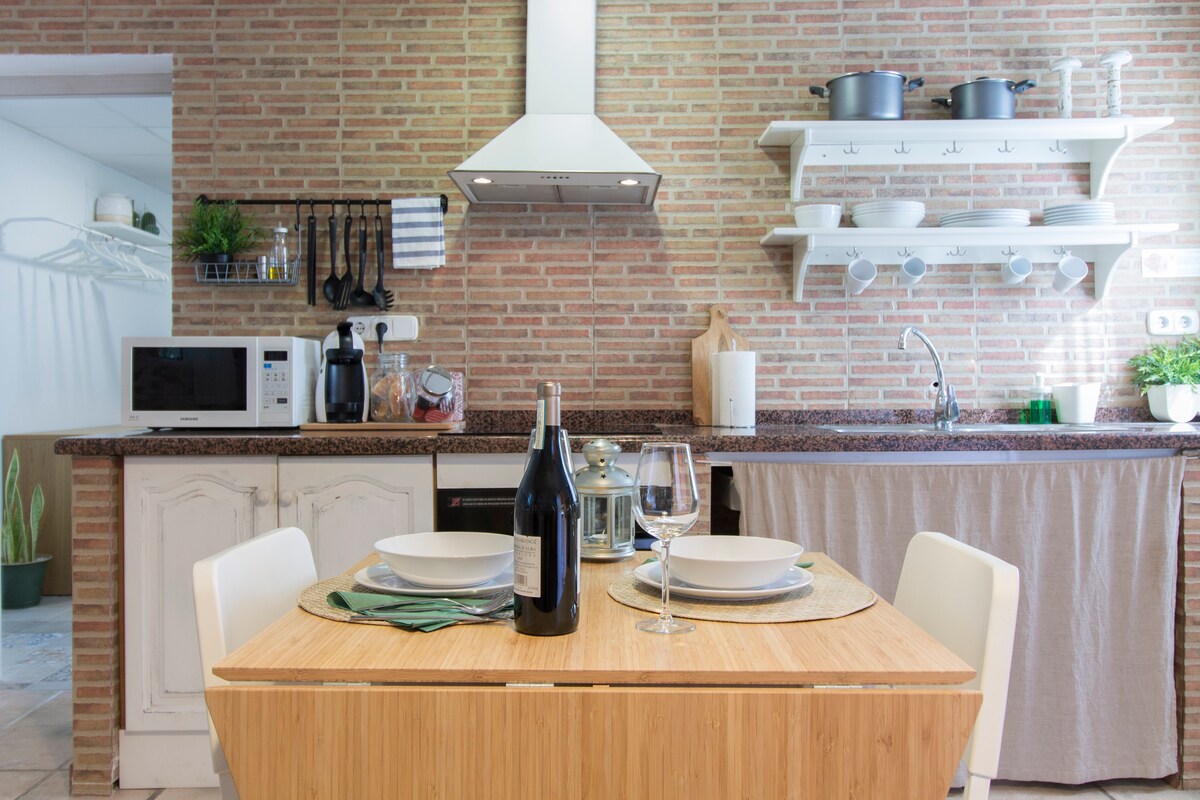 A modern kitchen is highlighted by a wooden table set for two, featuring plates and wine glasses. Brick-patterned walls complement stainless steel appliances and open shelving with various kitchen items. A microwave and additional kitchenware are visible, along with greenery for a touch of freshness.