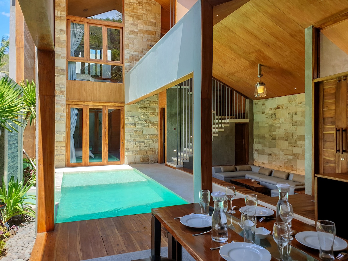 An open-plan living space features a dining area with a wooden table set for six, complemented by elegant glassware. Natural light floods the area from large windows, highlighting a private pool just beyond. Contemporary seating creates a relaxed atmosphere within the room.