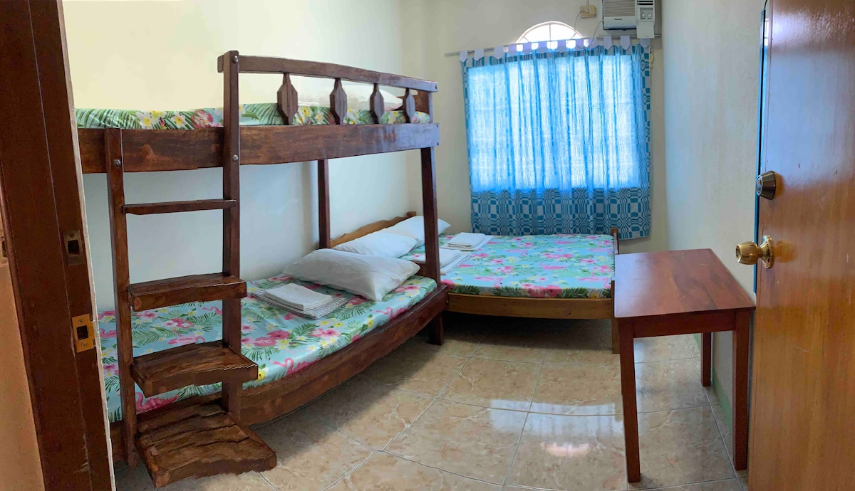 A simple room is furnished with a double size bed and a bunk bed, designed for comfort. Natural light filters through a set of windows adorned with light blue curtains. A wooden table is positioned near the door, enhancing the room's functional layout.