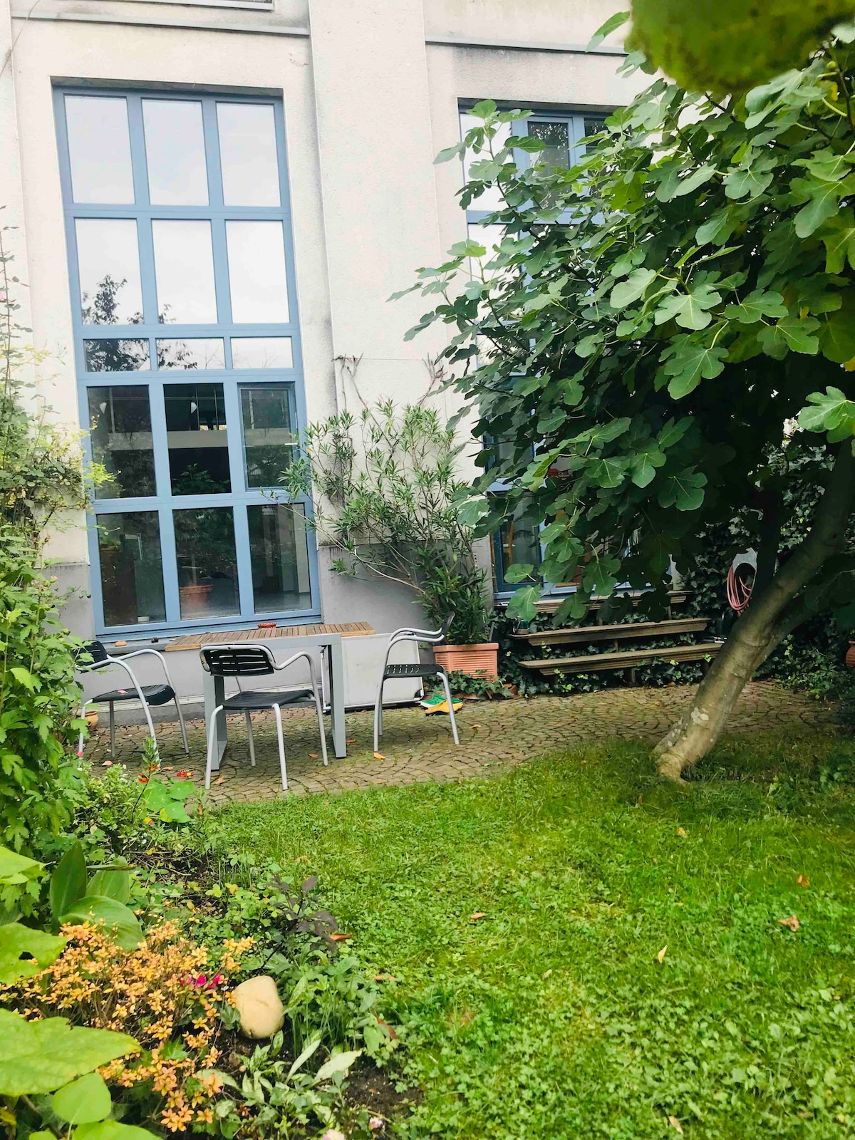 A peaceful garden area is framed by tall greenery, featuring a small table with three black chairs. Vibrant foliage and flowering plants enhance the outdoor space, while a wooden bench offers additional seating against a backdrop of large windows that allow natural light to filter in.