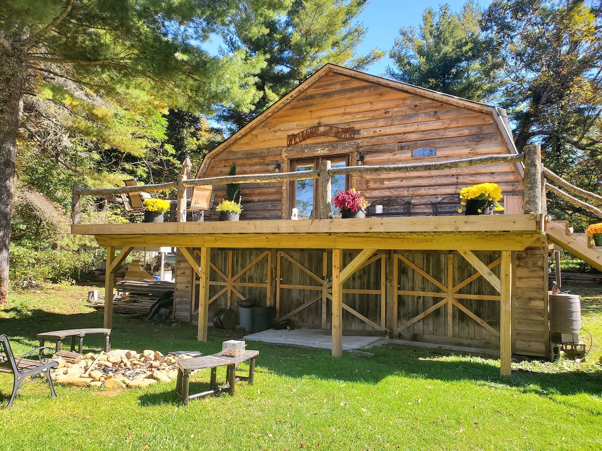 A charming wooden barn loft is set elevated on a grassy area, featuring a large deck adorned with potted flowers. Below, a fire pit area is visible with scattered logs and chairs, surrounded by tall trees that provide a natural backdrop.