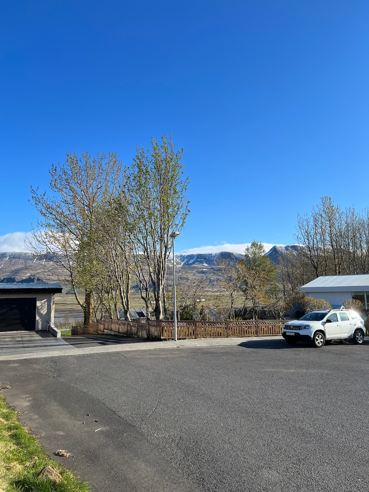 A view of the neighborhood is presented, featuring a quiet street lined with trees. In the background, mountains are visible under a clear blue sky. A parked car is situated in the foreground, with a garage and a sheltered area nearby.