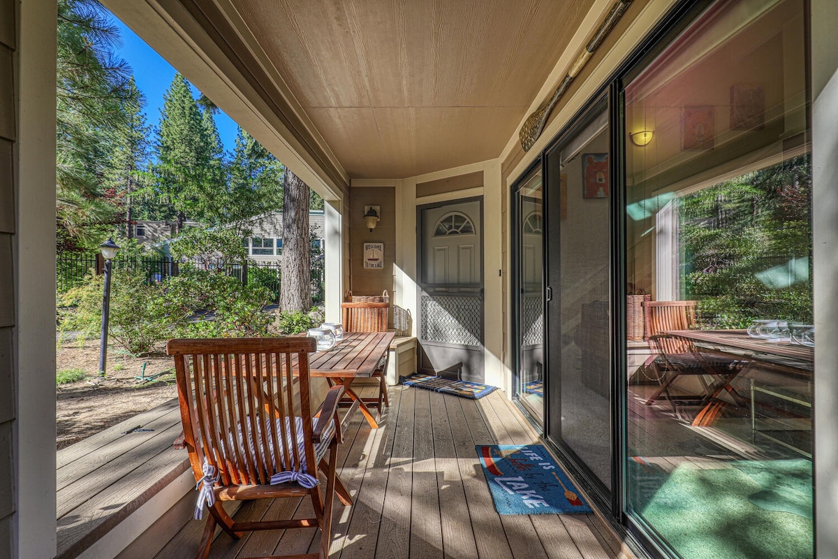 An inviting front patio area is visible, featuring wooden furniture including a table and chairs. Sliding glass doors lead into the interior, reflecting the greenery outside. A welcome mat is positioned at the entrance, enhancing accessibility and comfort.