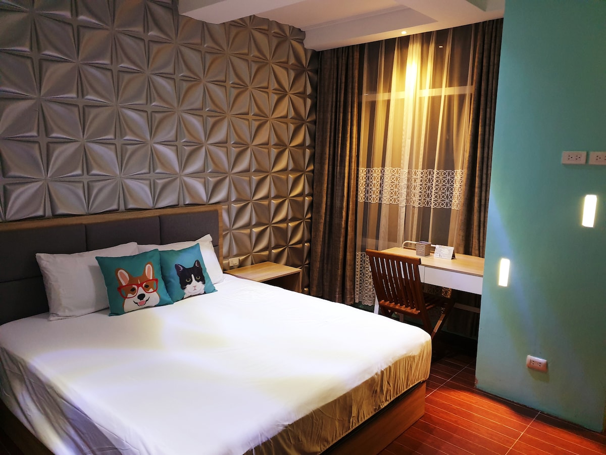 A spacious bedroom features a large bed with decorative throw pillows showcasing playful animal faces. A textured accent wall enhances the modern design, while large windows dressed with sheer curtains allow natural light to fill the room. A small desk and chair are also visible.