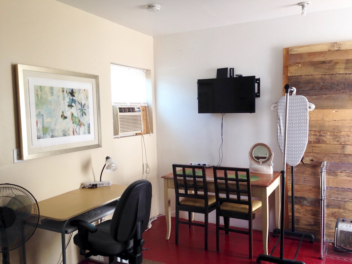 The studio room features a compact working area with a computer desk and two chairs. A wall-mounted television is positioned nearby, and a vintage ironing board stands against a wood-paneled accent wall. A window unit air conditioner provides climate control, while a standing fan offers additional air circulation.