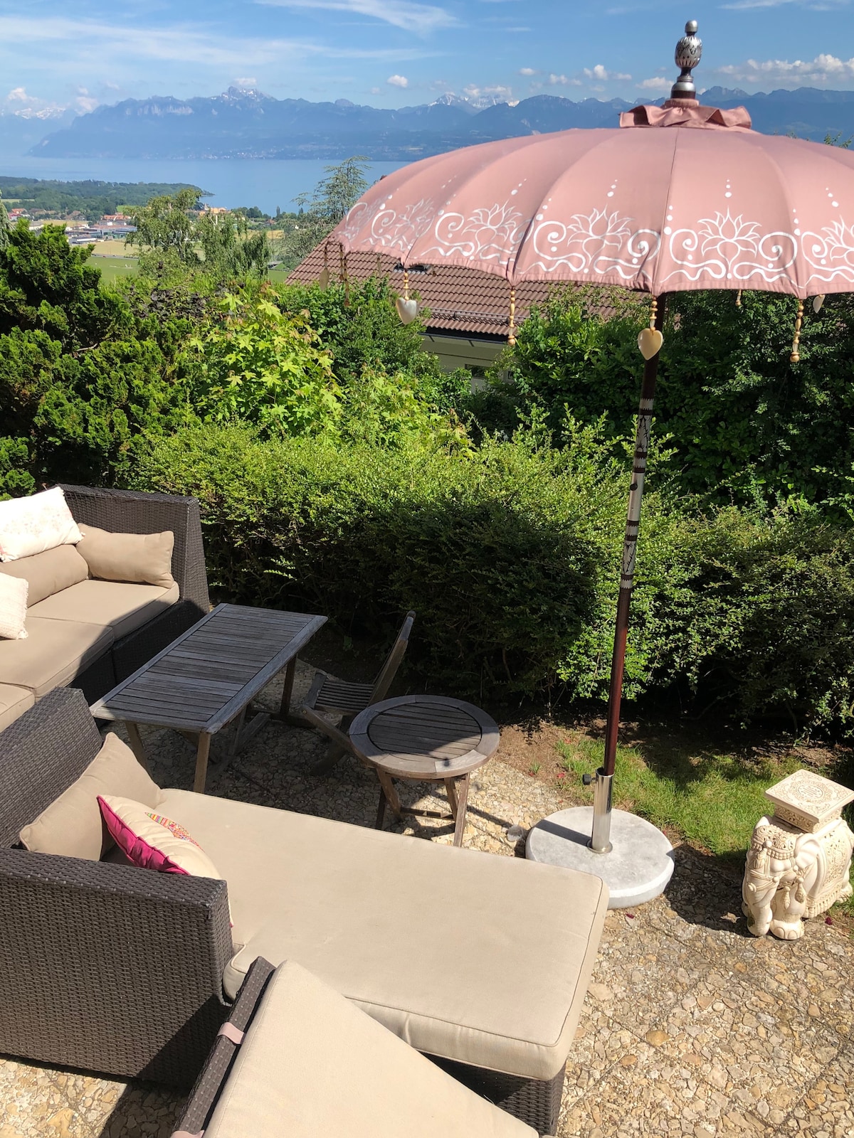 A private terrace features a comfortable seating area with a sectional sofa and armchair, complemented by a small table. A decorative umbrella provides shade, while greenery surrounds the space. A distant view of the mountains and the lake adds to the serene environment.