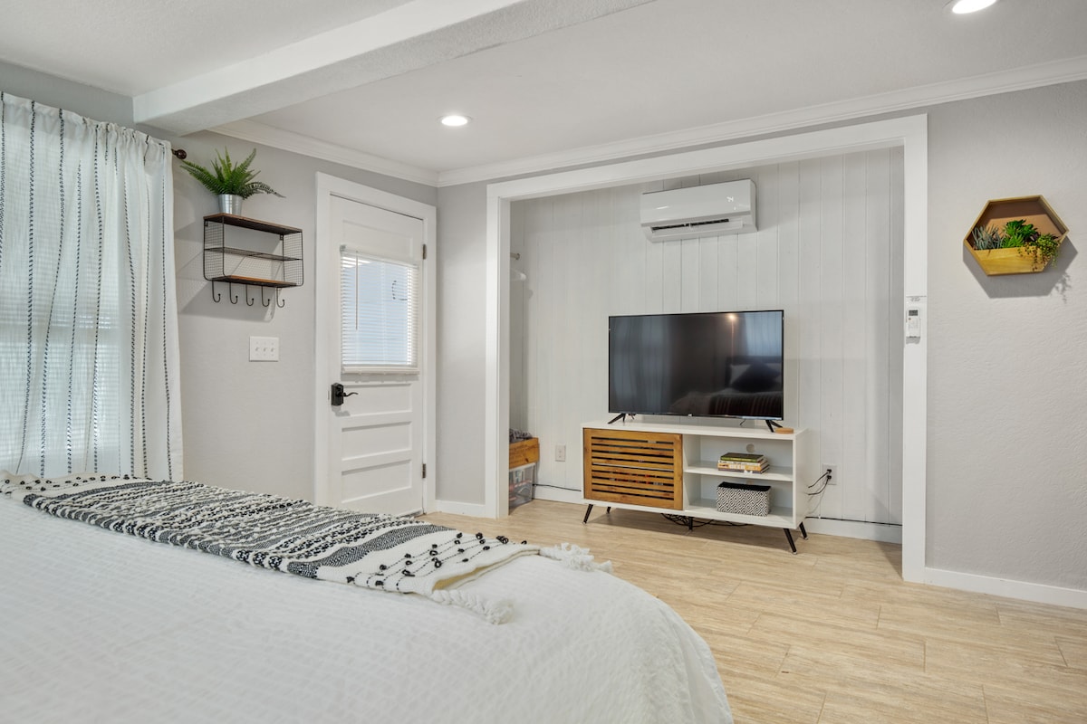 A modern room features a spacious bed adorned with a textured throw, adjacent to a TV mounted on a minimalist entertainment unit. Natural light flows through sheer curtains, and decorative elements include a plant on a shelf and a geometric wall planter.