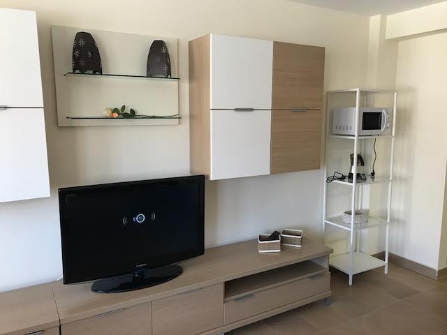 A modern living area features a flat-screen television placed on a light wood entertainment unit. Above the unit, stylish cabinets with a mix of white and wood finishes provide storage. A small microwave is positioned on a metallic stand to the right.
