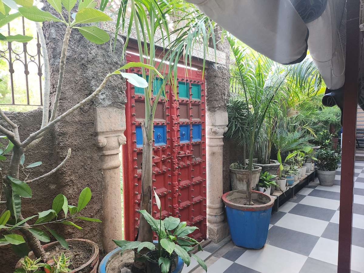A vibrant red door with intricate designs stands prominently against a textured stone wall. Lush green plants are arranged in various pots, creating a serene atmosphere. The pathway features alternating black and white tiles, leading towards the door, surrounded by a canopy that provides shade.
